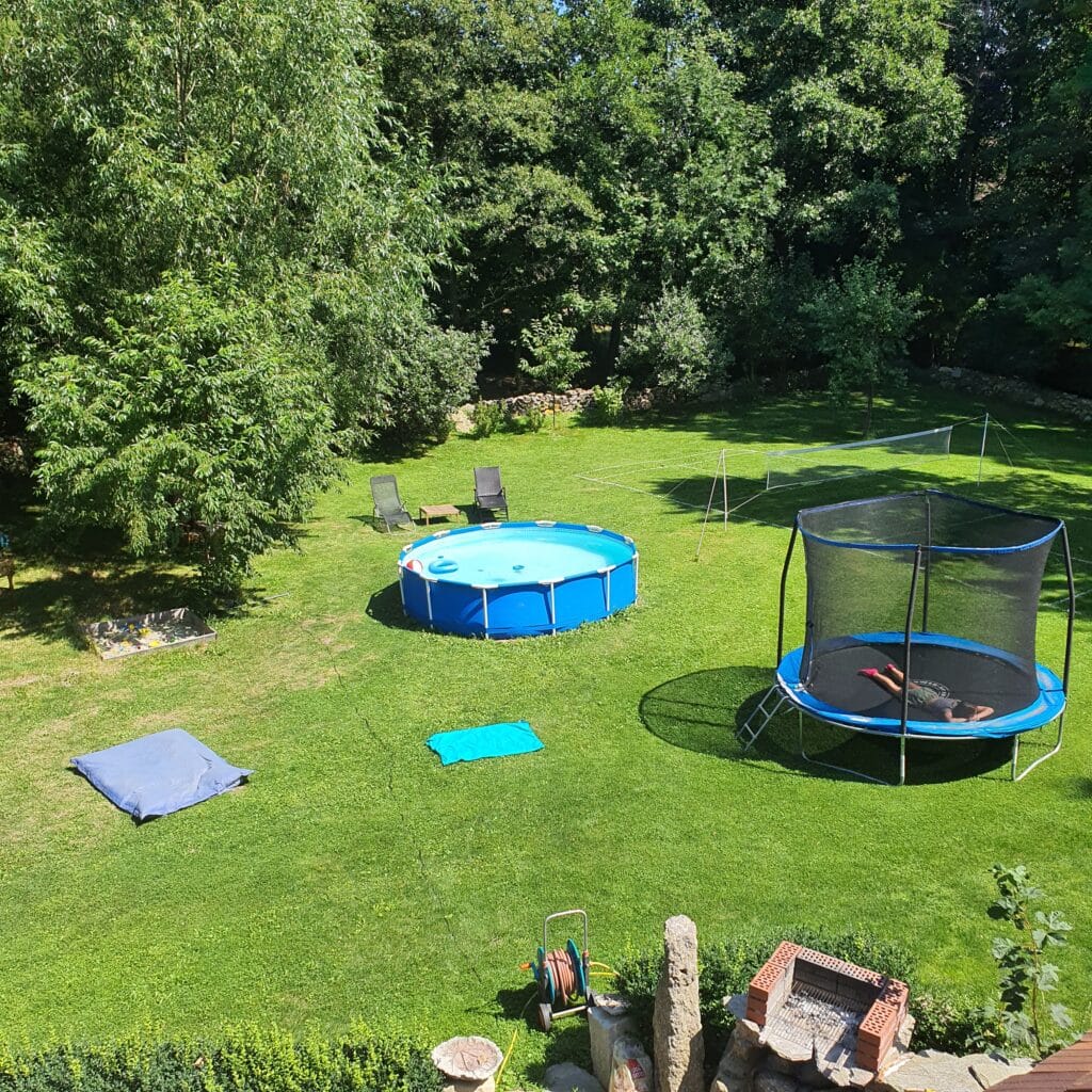 20230815_125006 - Dworek Marmis Zielony ogród z basenem, trampoliną, leżakami i kocami na trawie. Drzewa w tle.