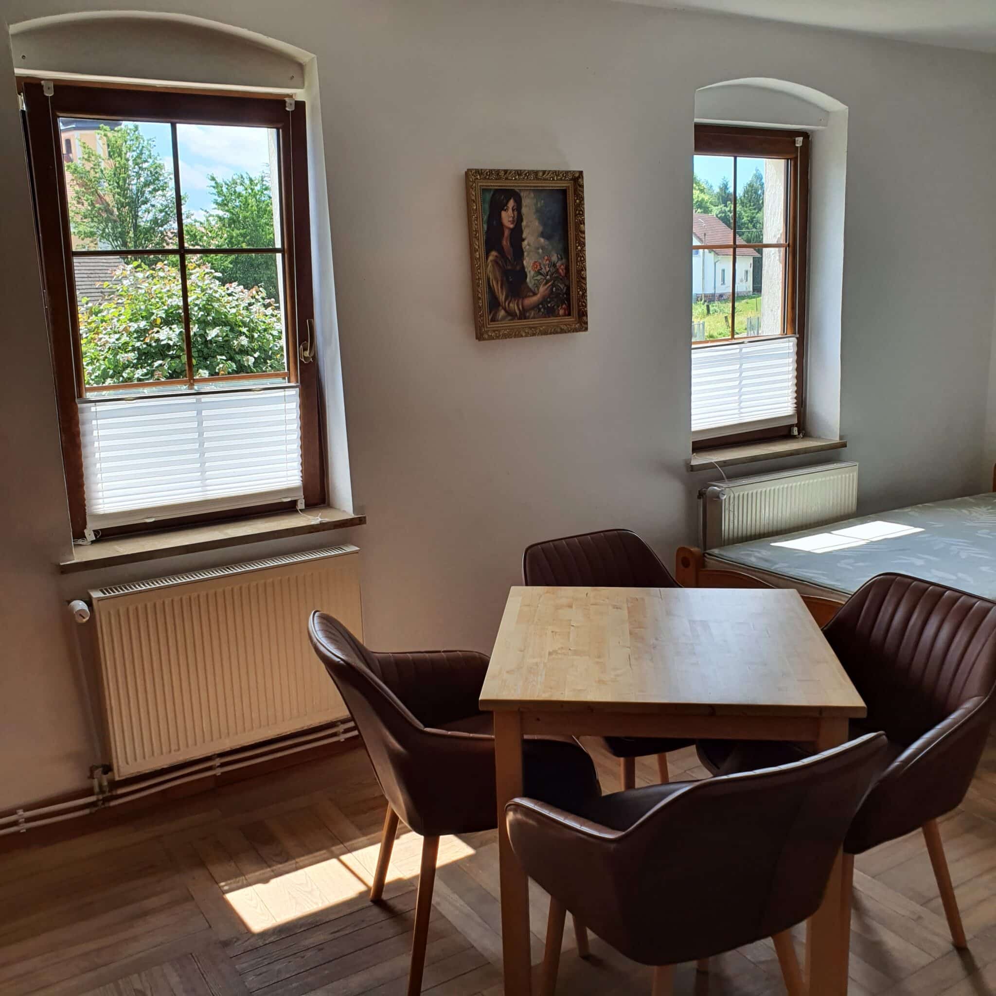 20250628_140240 - Dworek Marmis Jasny pokój kominkowy z drewnianym stołem i czterema krzesłami, widok na okna z zielonymi drzewami.