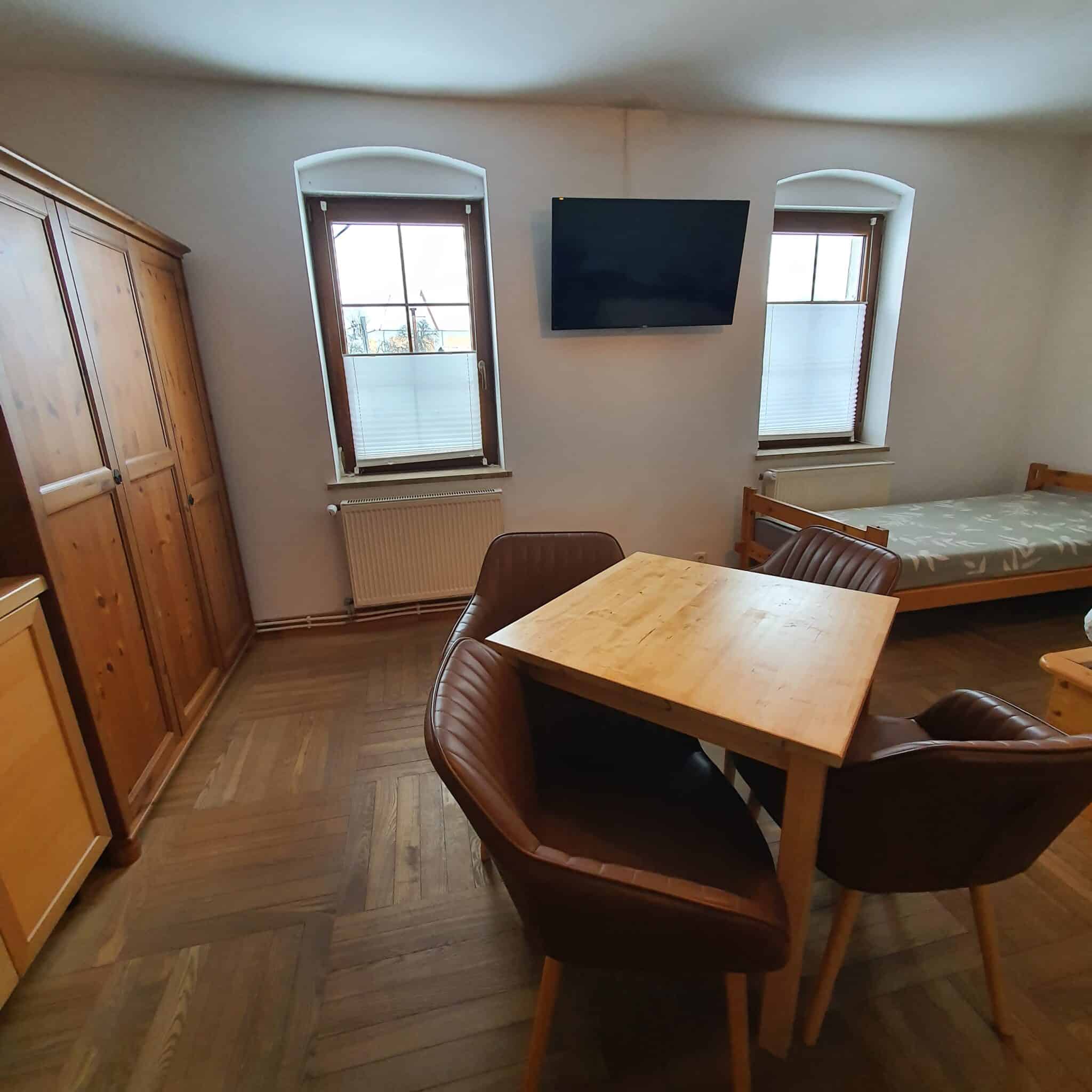 20260130_100128 - Dworek Marmis Pokój kominkowy z drewnianym stołem i krzesłami, szafą oraz łóżkiem w tle.