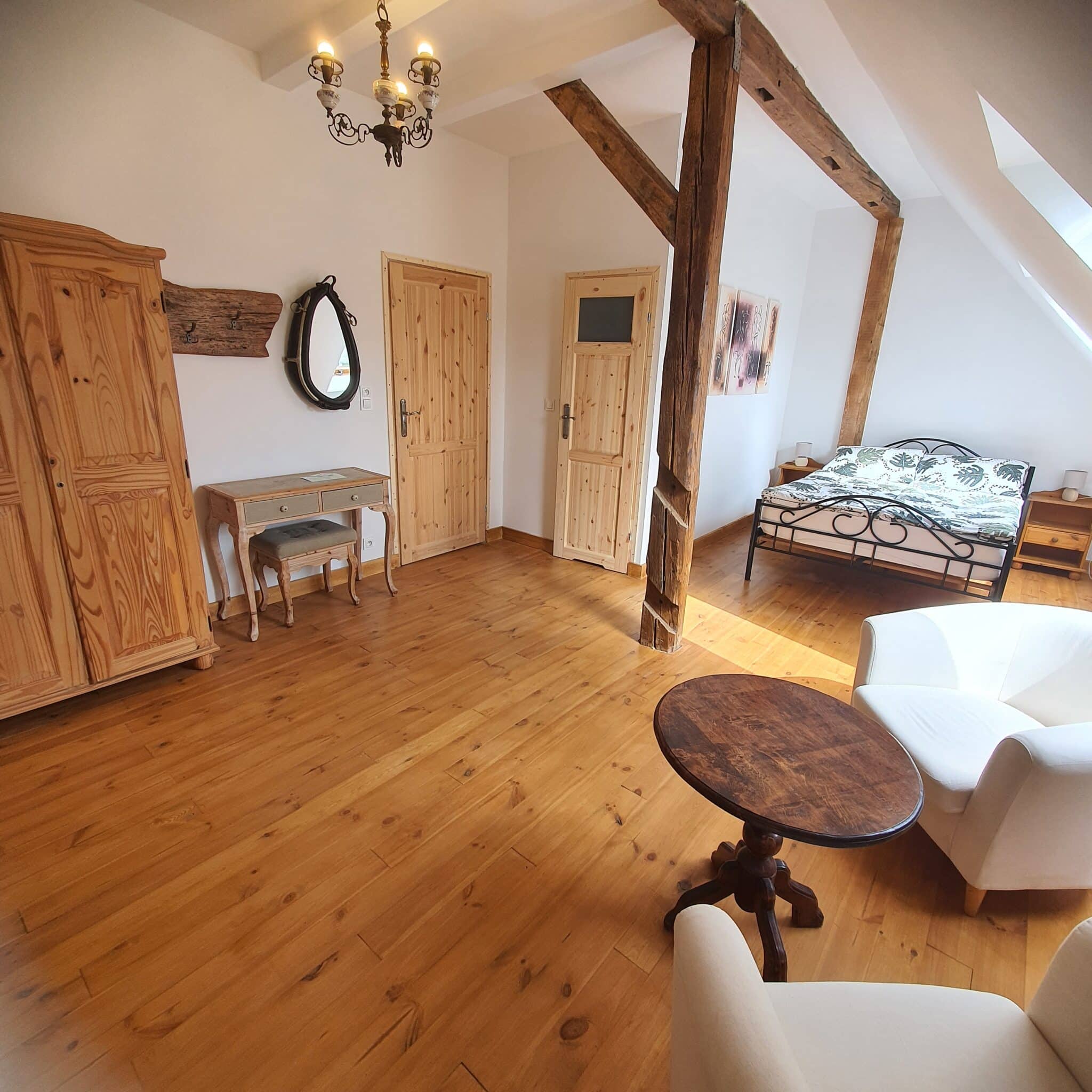 Przestronny pokój z drewnianą podłogą, łóżkiem, szafą, lustrem, stolikiem i białym fotelem.