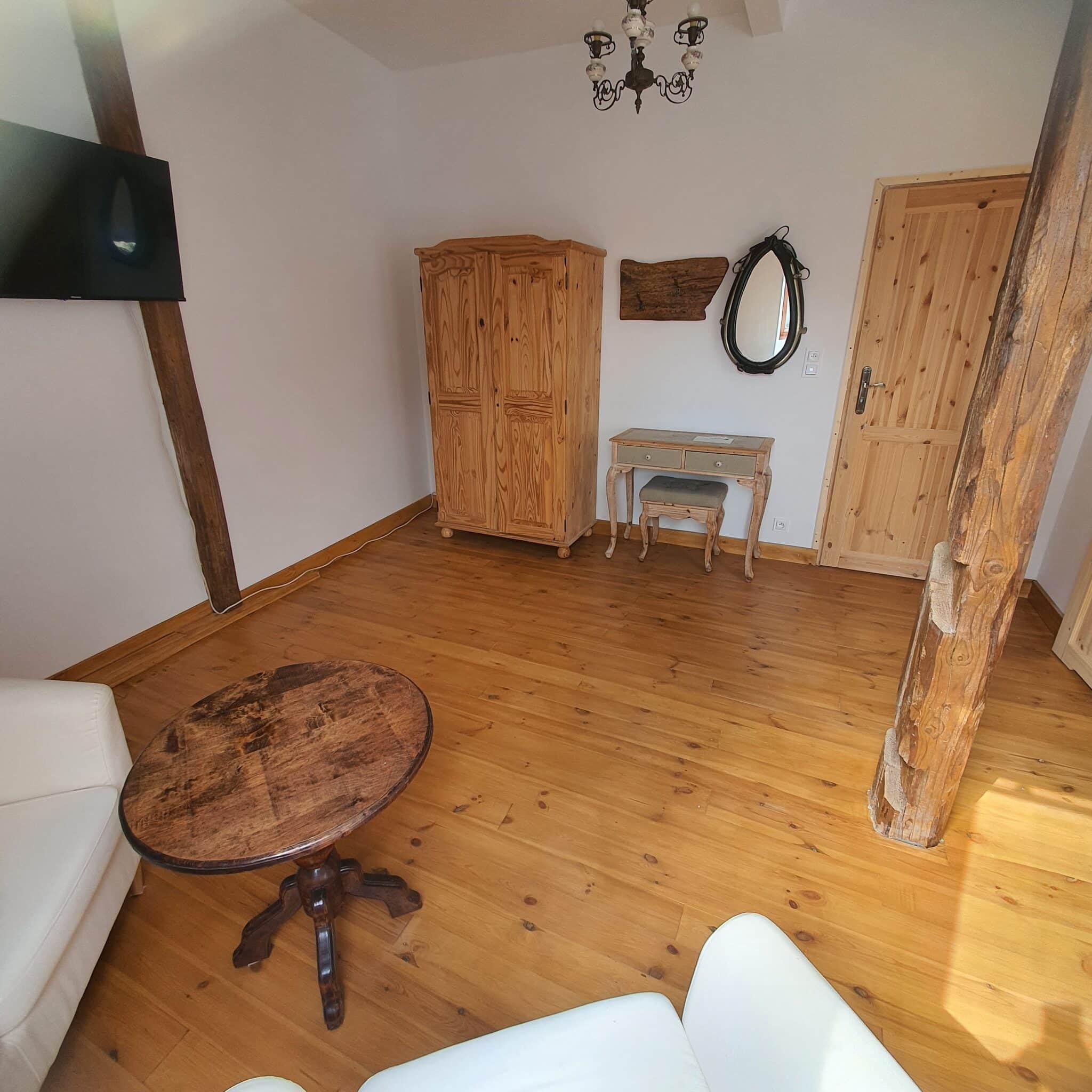 20250717_155210 - Dworek Marmis Wnętrze pokoju z drewnianą podłogą, białymi sofami, stolikiem kawowym i szafą. Na ścianie telewizor i lustro.