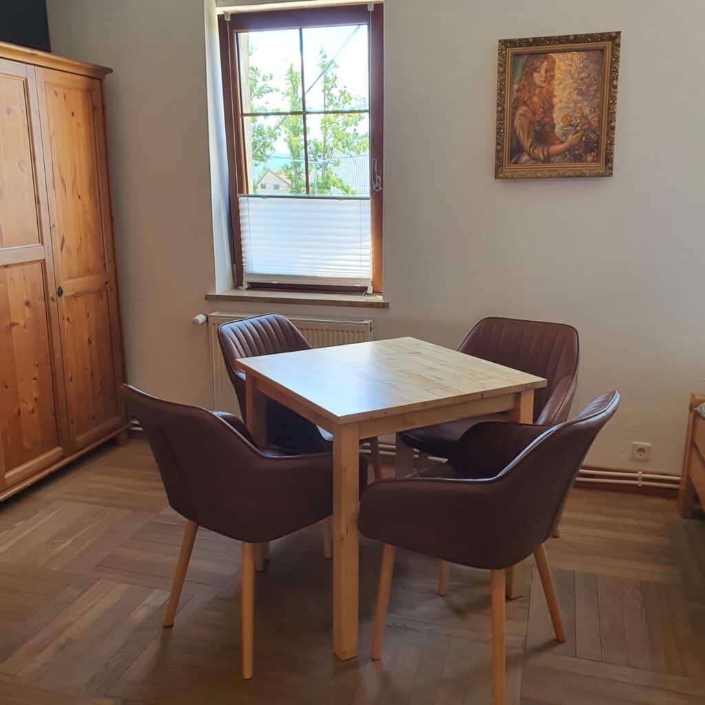 20250619_121451 - Dworek Marmis Heller Holztisch umgeben von vier braunen Stühlen in einem hellen Raum. Im Hintergrund ein Fenster und ein Gemälde an der Wand.