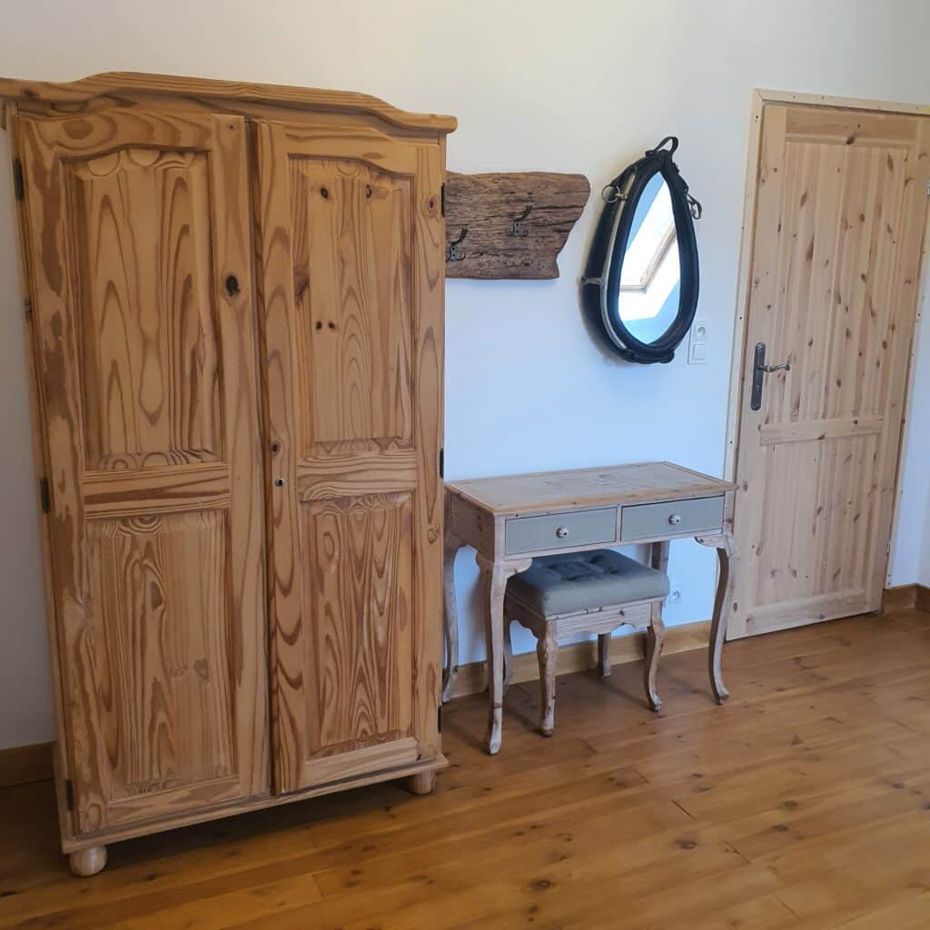 20250717_144144 - Dworek Marmis Ein hölzerner Kleiderschrank, ein Tisch mit Schubladen und ein gerahmter Spiegel vor einer hellen Wand und einem Holzboden.