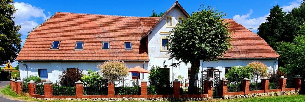 20250723_142914 - Dworek Marmis Biały dom z czerwoną dachówką, otoczony zielenią, z ogrodzeniem z cegły i drzewem przed wejściem.