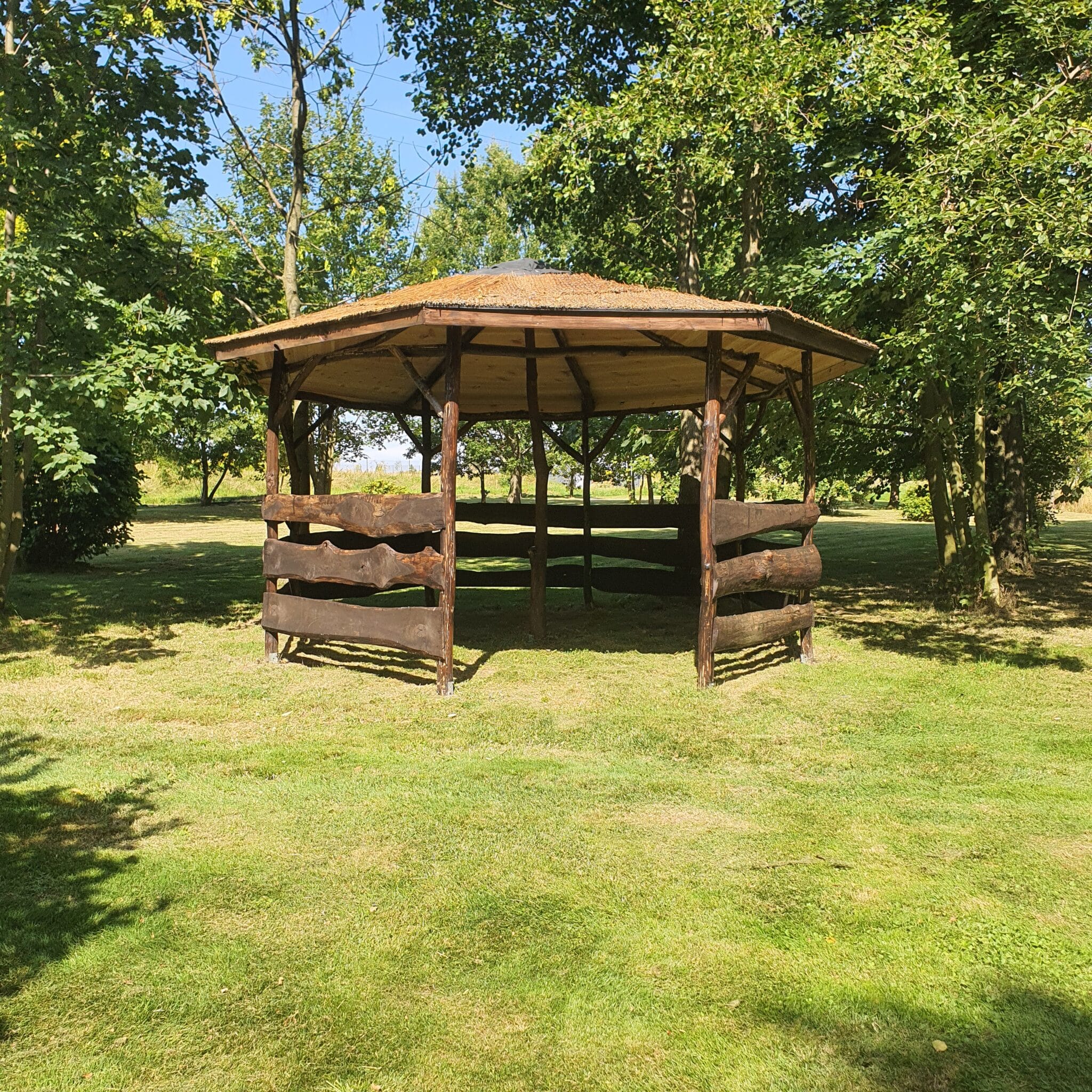 20250828_143257 - Dworek Marmis Alt: Altana z drewnianymi ścianami i dachem, otoczona zielonym trawnikiem i drzewami w Dworku Marmis.