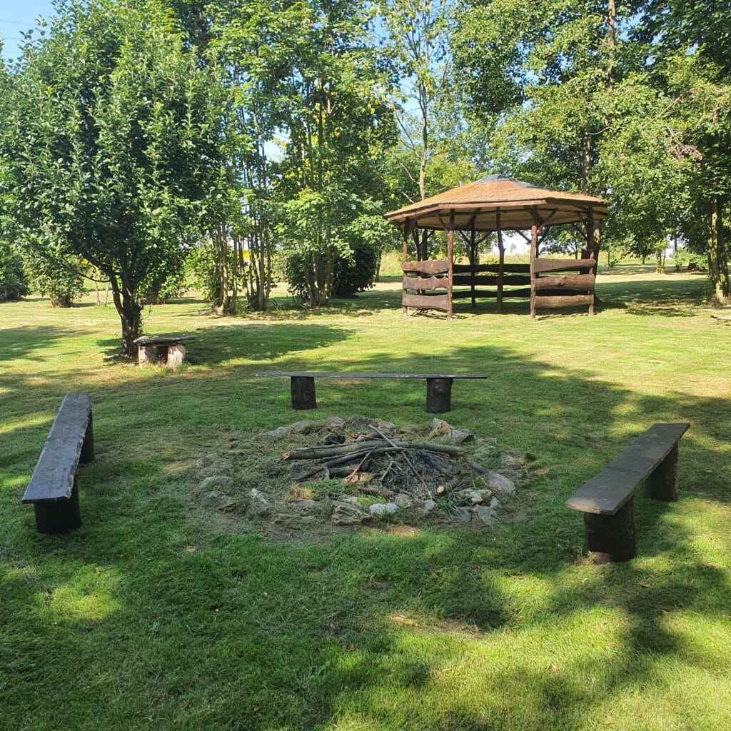 20250828_143323 - Dworek Marmis Ein Lagerfeuer, umgeben von Holzbänken in einer grünen Umgebung, mit einem Pavillon im Hintergrund.