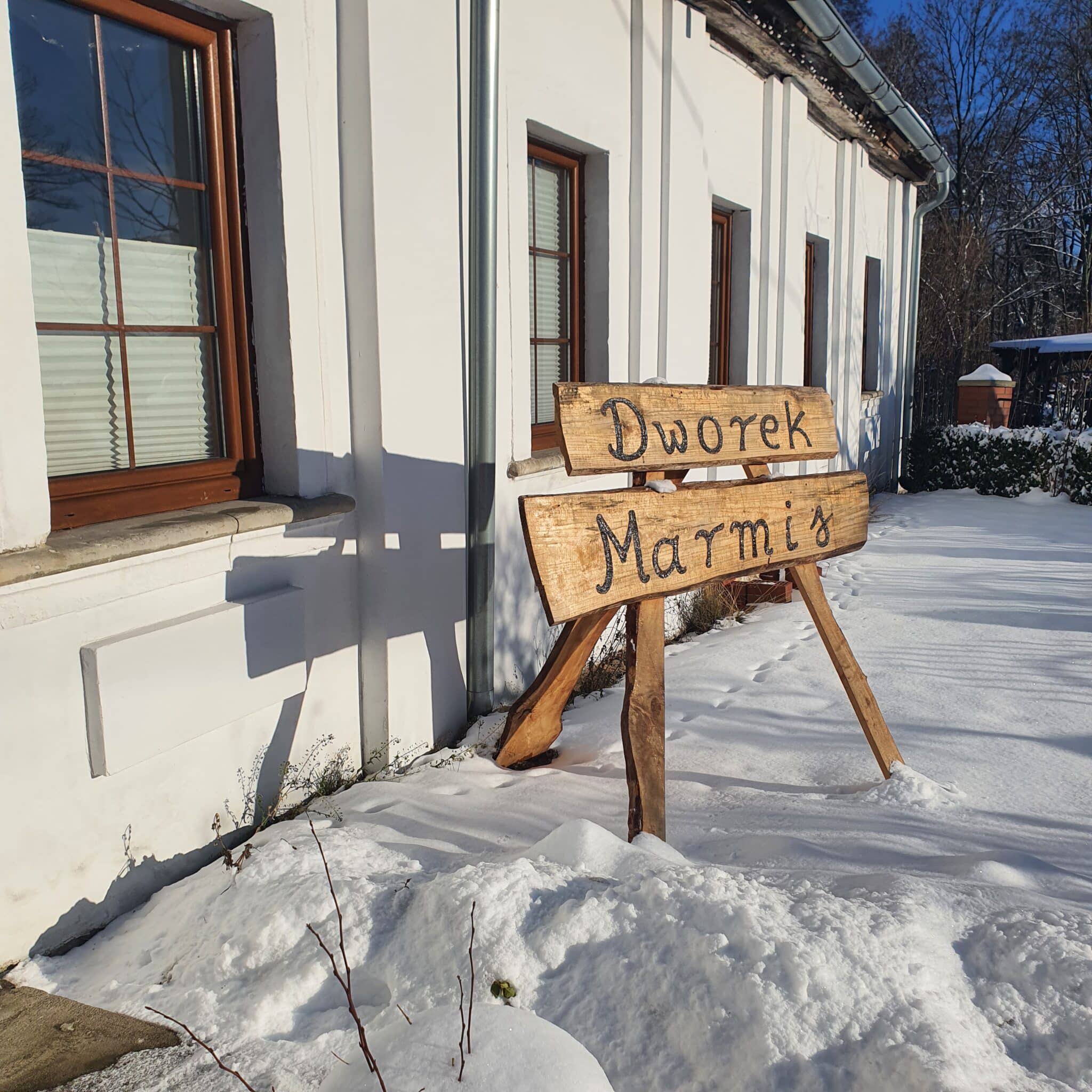 20260110_135654 - Dworek Marmis Dworek Marmis, dřevěná cedule stojící ve sněhu vedle bílé budovy.