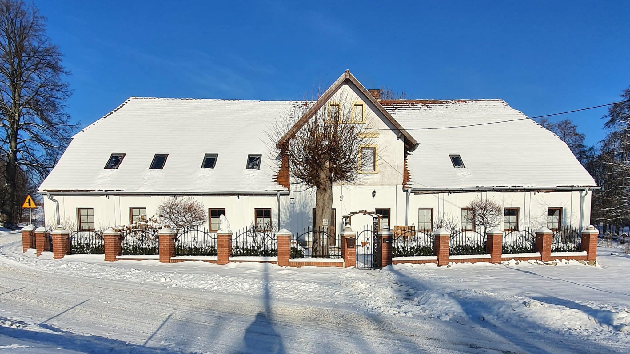 20260110_140728 - Dworek Marmis Dworek Marmis, biały budynek pokryty śniegiem, z ogrodzeniem i drzewami, w słoneczny dzień.