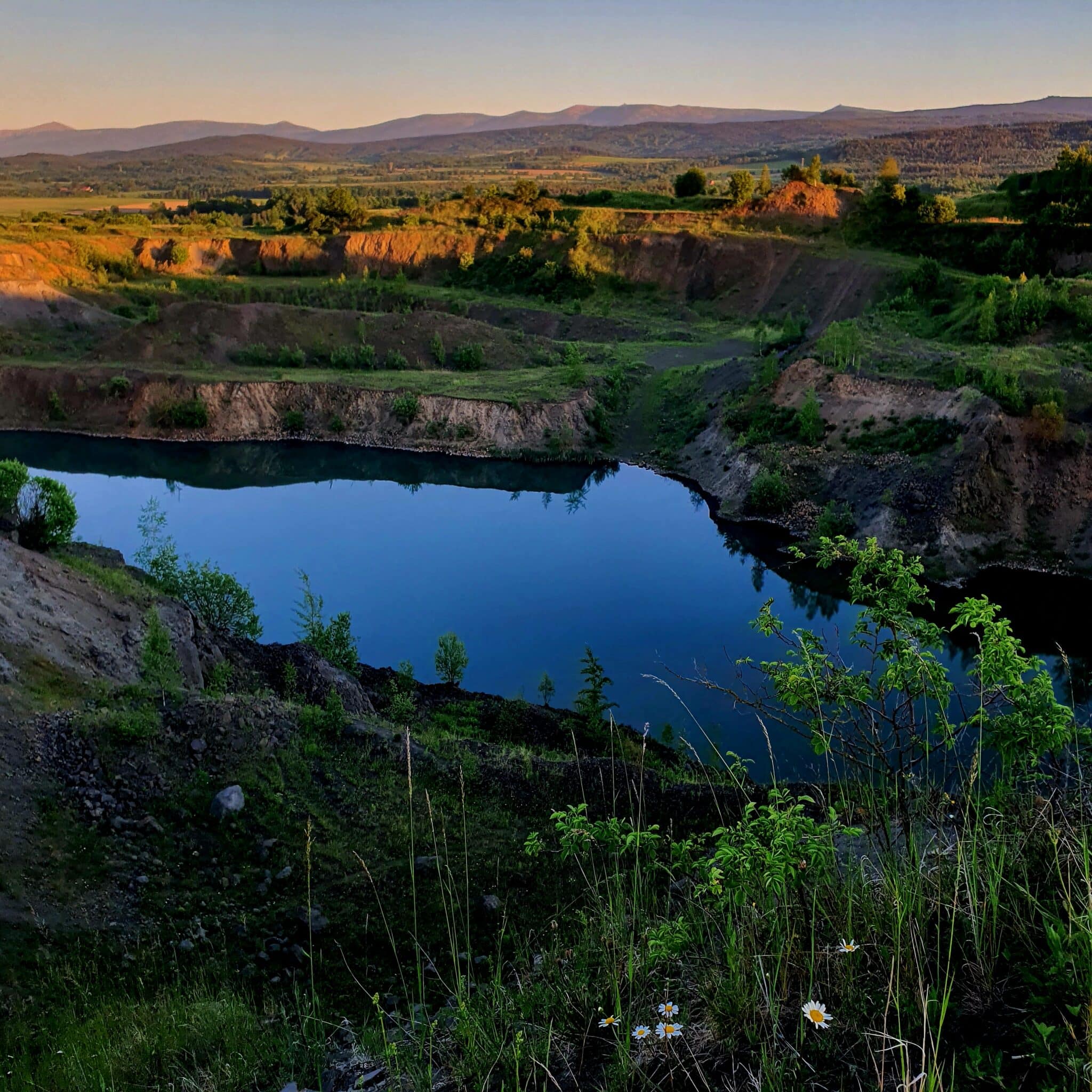 20260525_202436 - Dworek Marmis Ein Blick auf einen ruhigen Teich, umgeben von grünen Hügeln und Blumen, mit Bergen im Sonnenuntergang im Hintergrund.