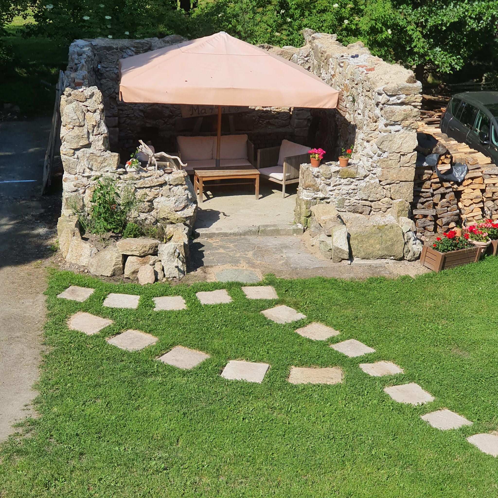 20260627_161758 – Dworek Marmis Zelená zahrada s kamennou terasou, slunečníkem a zahradním nábytkem, obklopená květinami.