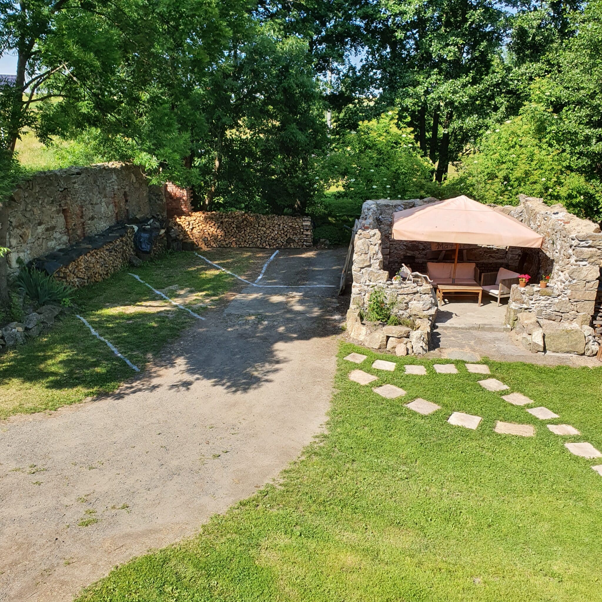 20260627_161819 – Dworek Marmis Pohled na zahradu z terasy, s kamennými zříceninami, dřevěným stolem a slunečníkem, obklopenou zelení.